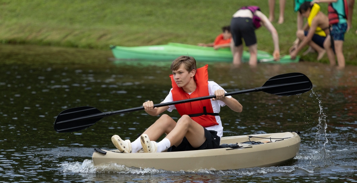 Image shows a student paddling a kayak in a lake. More students are standing on the shore.