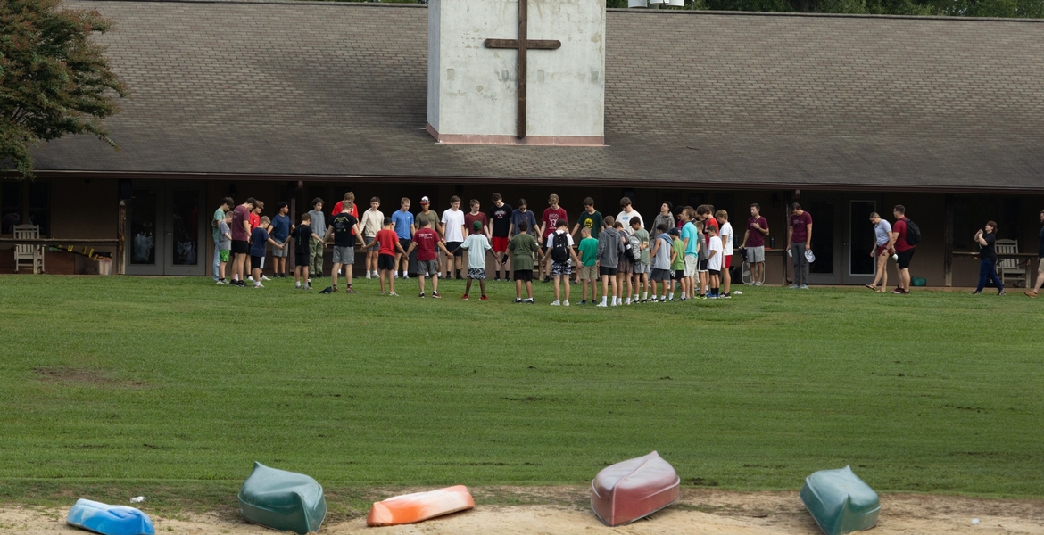 Image shows a group of students & teachers standing in a circle praying outside.