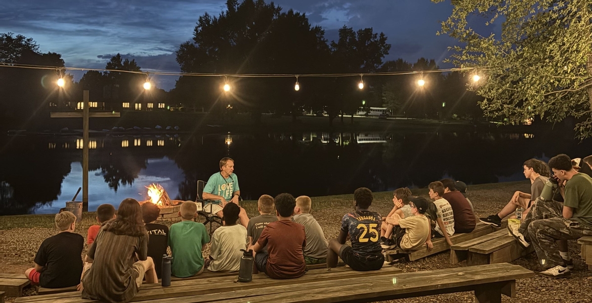 Image shows students sitting near a lake at dusk listening to teaching from the head of school.