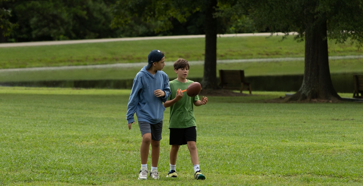 Image shows an older student walking across a field with a younger student carrying a football.